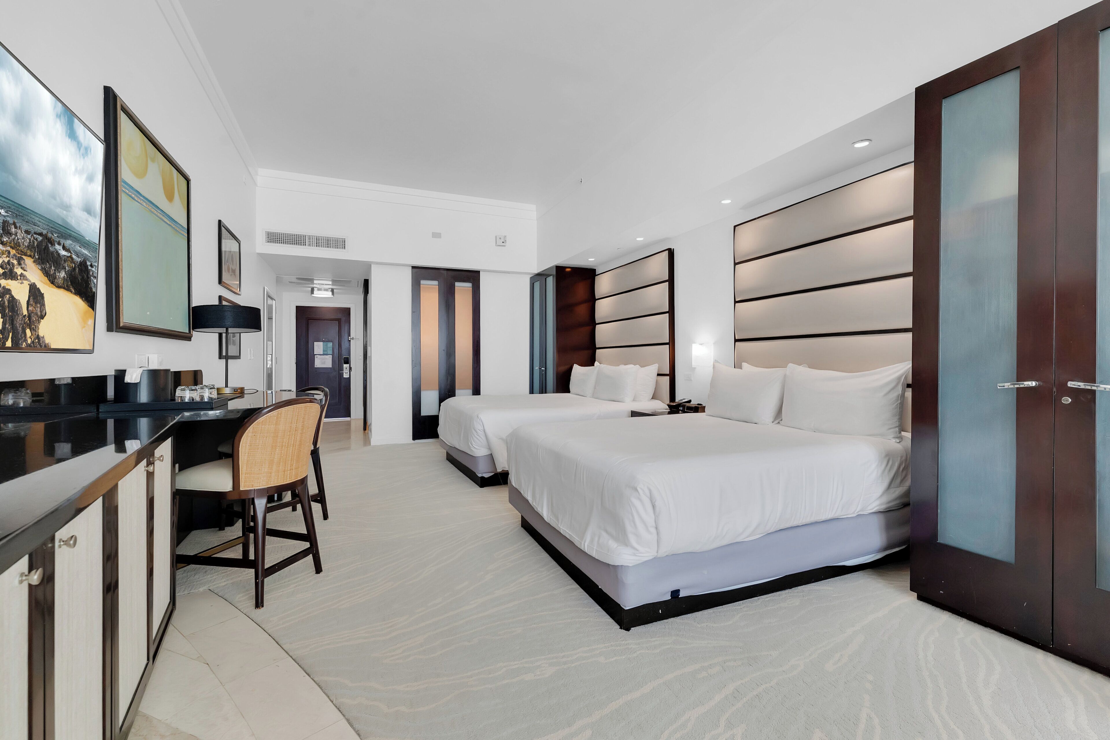 A hotel room with two beds, a desk, and a chair. There is a large painting on the wall and a wardrobe with a mirror.