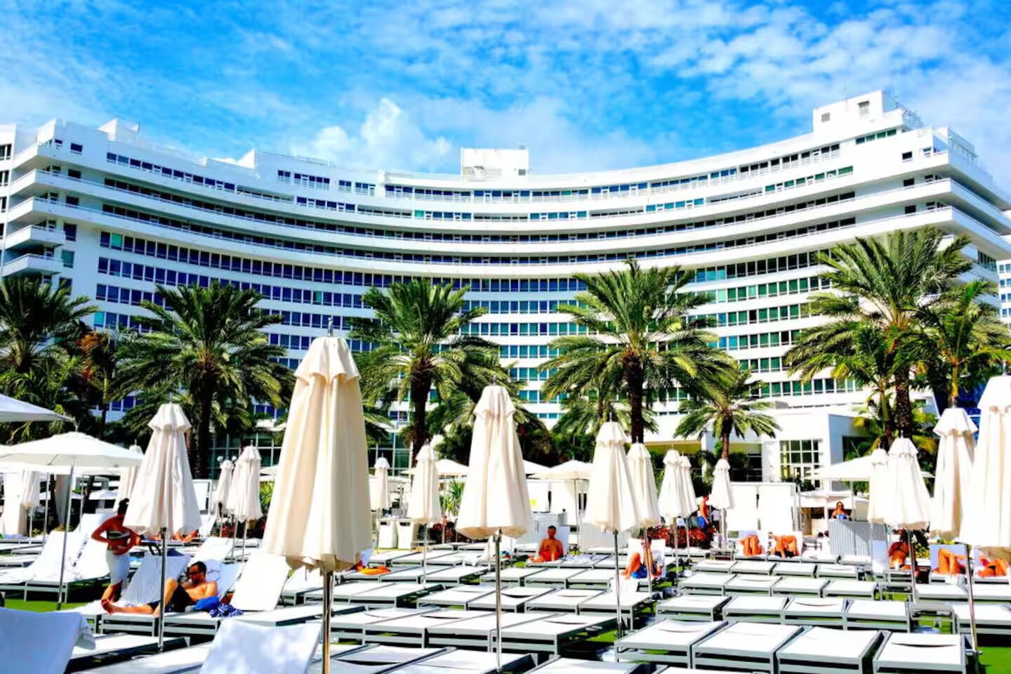 Um hotel com fachada curva, várias varandas e área de piscina com espreguiçadeiras e guarda-sóis.
