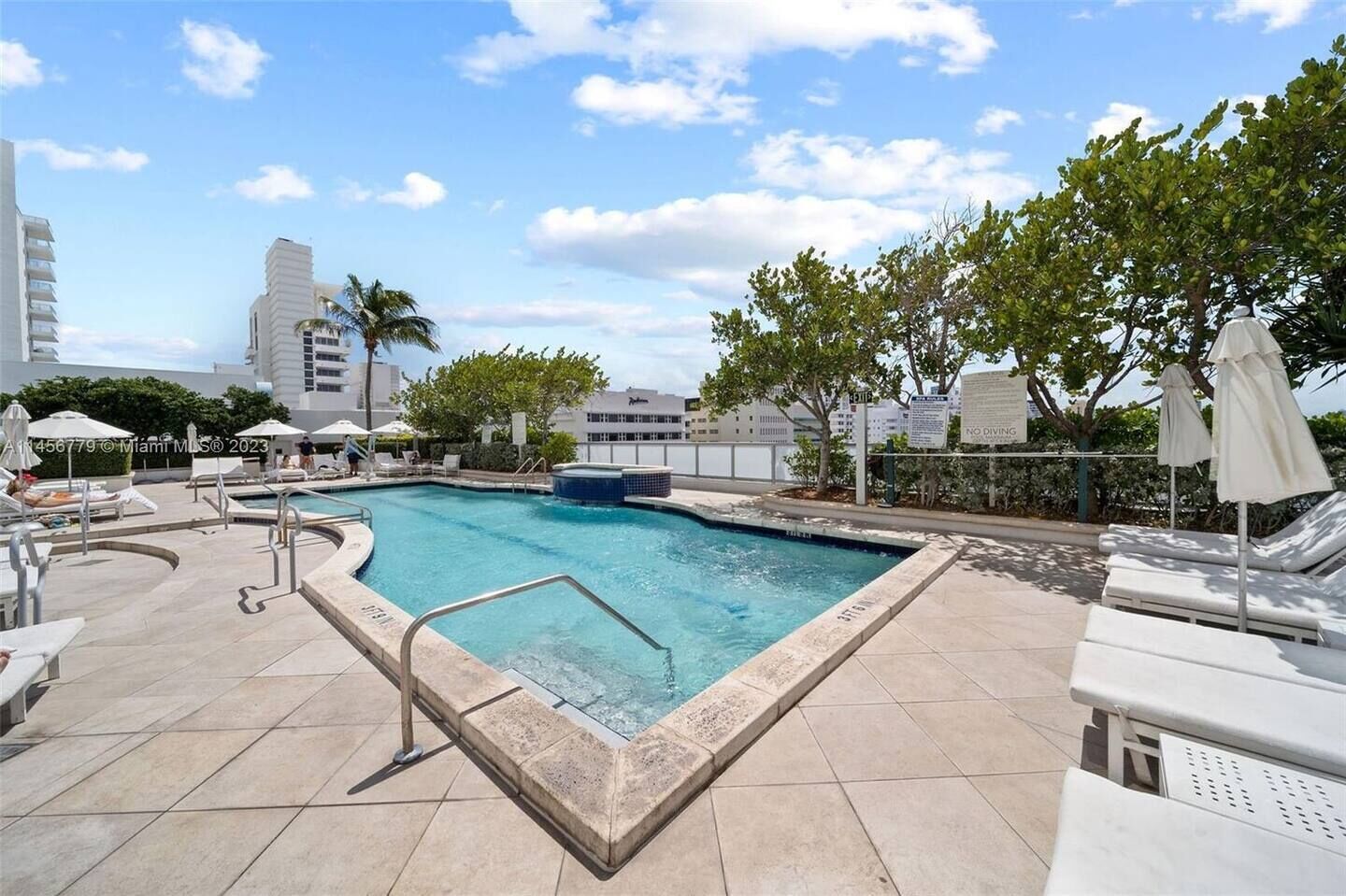 A swimming pool with lounge chairs and umbrellas, surrounded by buildings and trees.