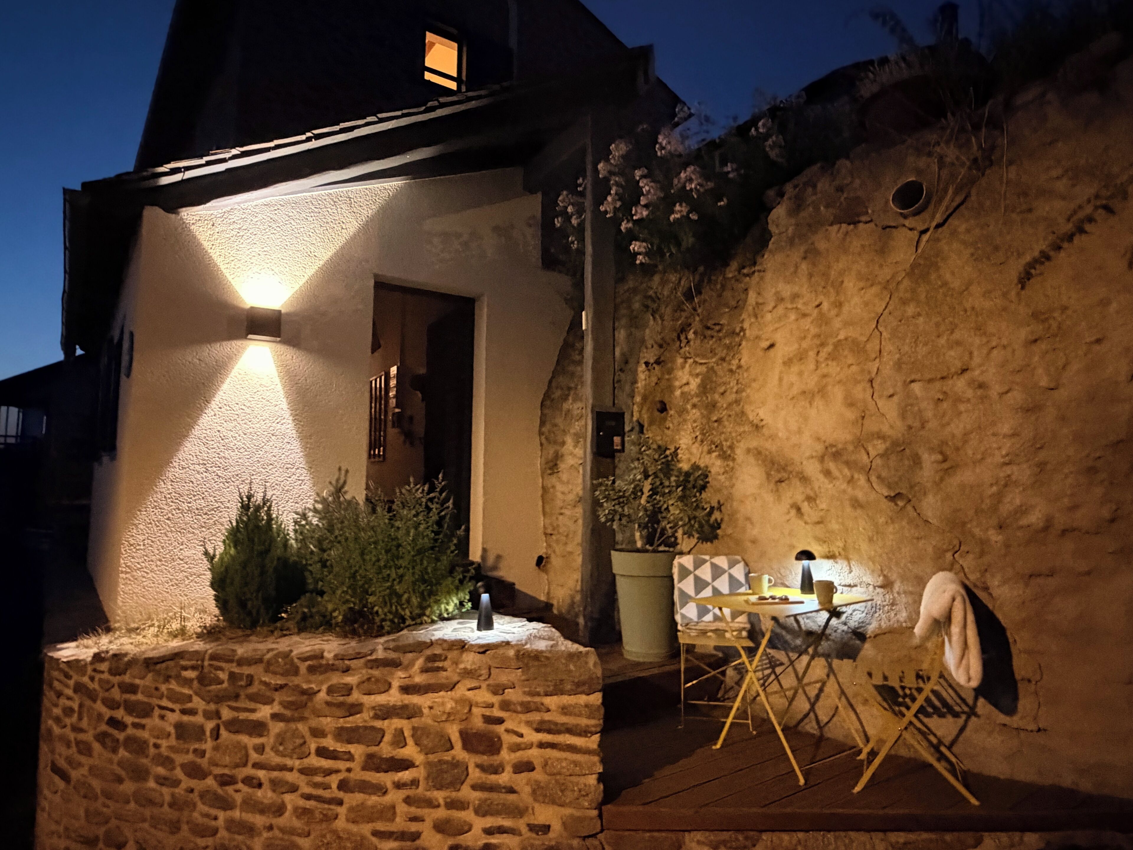 Un muro de piedra con una pequeña mesa y sillas, una planta en maceta y una ventana con luz encendida.
