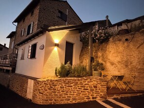 A stone building with a sloped roof, a lit exterior wall, and a small garden area with a bench.