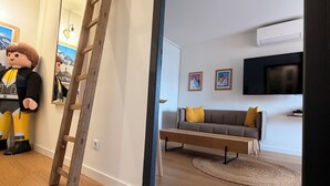 Una sala de estar moderna con piso de madera, un sofá con cojines, un televisor de pantalla plana y una escalera apoyada en la pared.