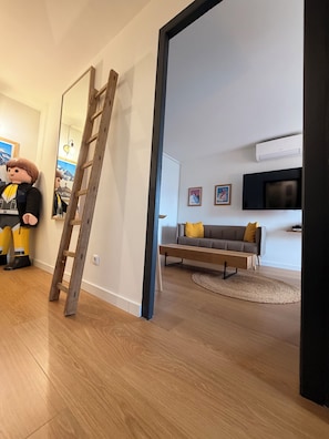 Uma sala de estar moderna com piso de madeira, um sofá com almofadas, uma televisão de tela plana e uma escada encostada na parede.
