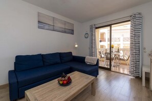 A living room with a blue sofa, a wooden coffee table with fruit, and a view of an outdoor balcony with chairs and a table. - Castle Harbour 42 - One Bed (Los Cristianos)