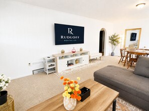 A modern living room with a flat-screen TV mounted on the wall, a wooden coffee table with a vase of flowers, a grey sofa, and a dining area with wooden chairs and a table. - The Overlook is a cozy two-bedroom retreat overlooking downtown Waynesville. Enjoy beautiful bluff views, a quiet neighborhood, and easy access to local restaurants, parks, shops, and Fort Leonard Wood. (Waynesville)