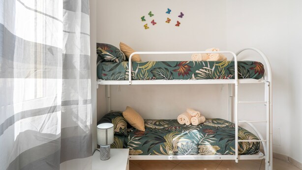 Una cama litera con estampado de hojas tropicales, una mesita de noche pequeña con lámpara y adhesivos de mariposas en la pared.