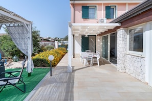 Un'area salotto esterna coperta con tavolo e sedie, un pergolato e vista su una strada residenziale.