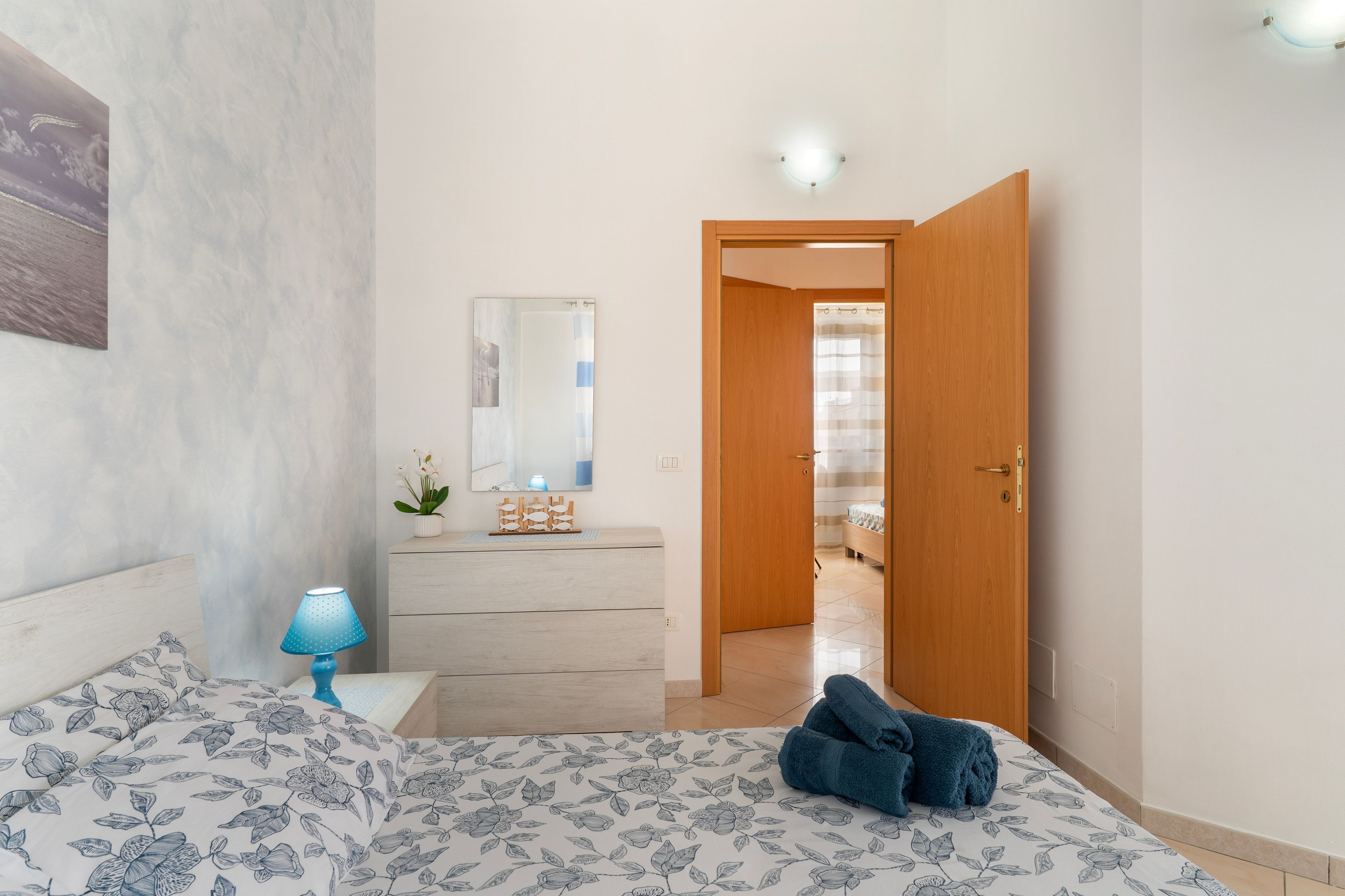 Ein Schlafzimmer mit Bett, Kommode, Nachttisch, Lampe und einer Tür zu einem anderen Raum.
