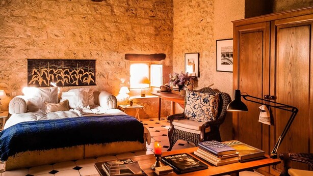 A rustic room with a bed, a chair, a table with books, and a lamp.