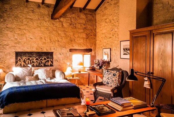 A rustic room with a bed, a chair, a table with books, and a lamp.