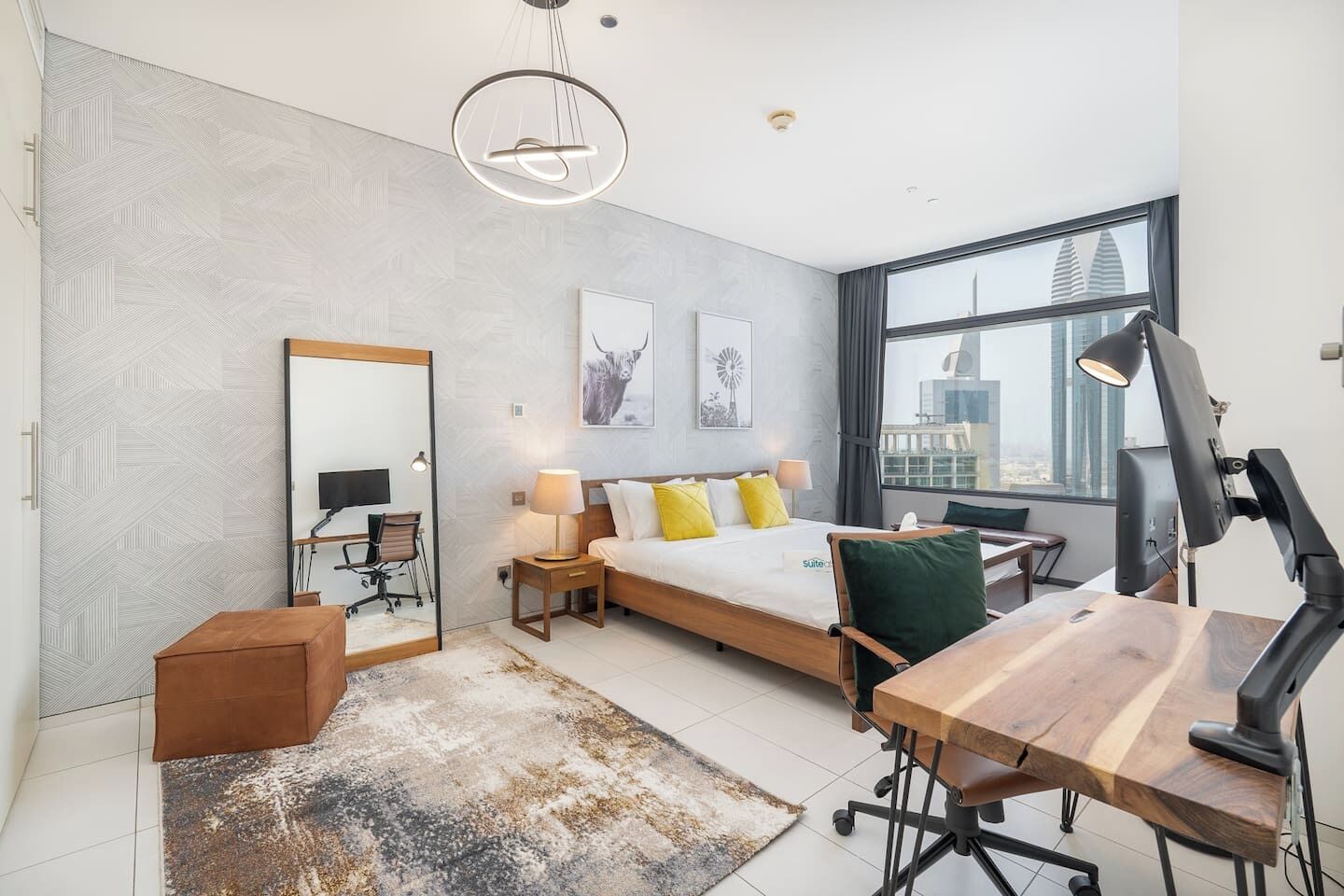 Et moderne hotelværelse med en stor seng, et skrivebord, en stol, en skrivebordslampe, et spejl og udsigt over byen gennem et stort vindue.