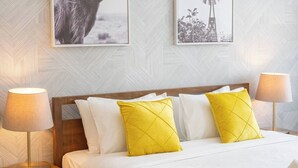 Hotellihuone, jossa on sänky, kaksi yöpöytälamppua, yöpöytä, jossa on yksi laatikko, ja kaksi kehystettyä kuvaa seinällä.