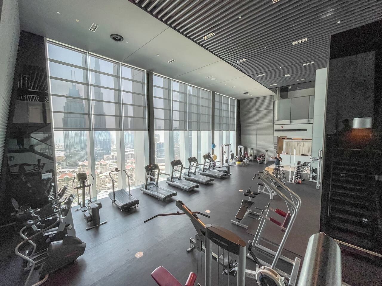 Een sportschool met loopbanden, hometrainers en krachtapparaten, grote ramen met stadszicht, en een modern plafondontwerp.