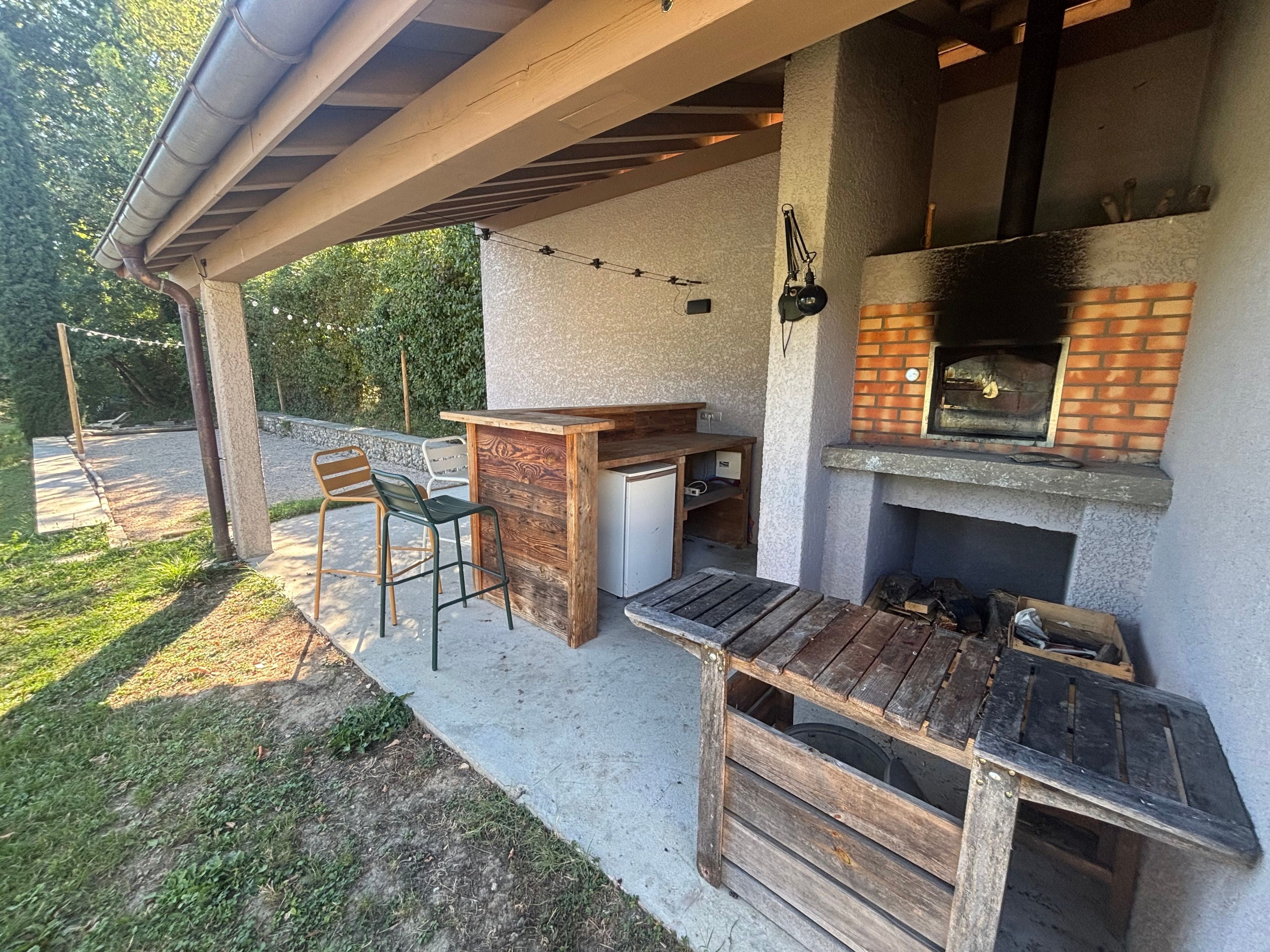 An outdoor patio with a wooden table and chairs, a built-in pizza oven, and a concrete floor.