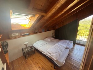 Un dormitorio con techo inclinado, una cama, una mesita de noche con lámpara, un pequeño estante con adornos y una ventana con vista a follaje.