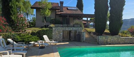 A pool area with lounge chairs, a stone wall, and a house with a tiled roof.