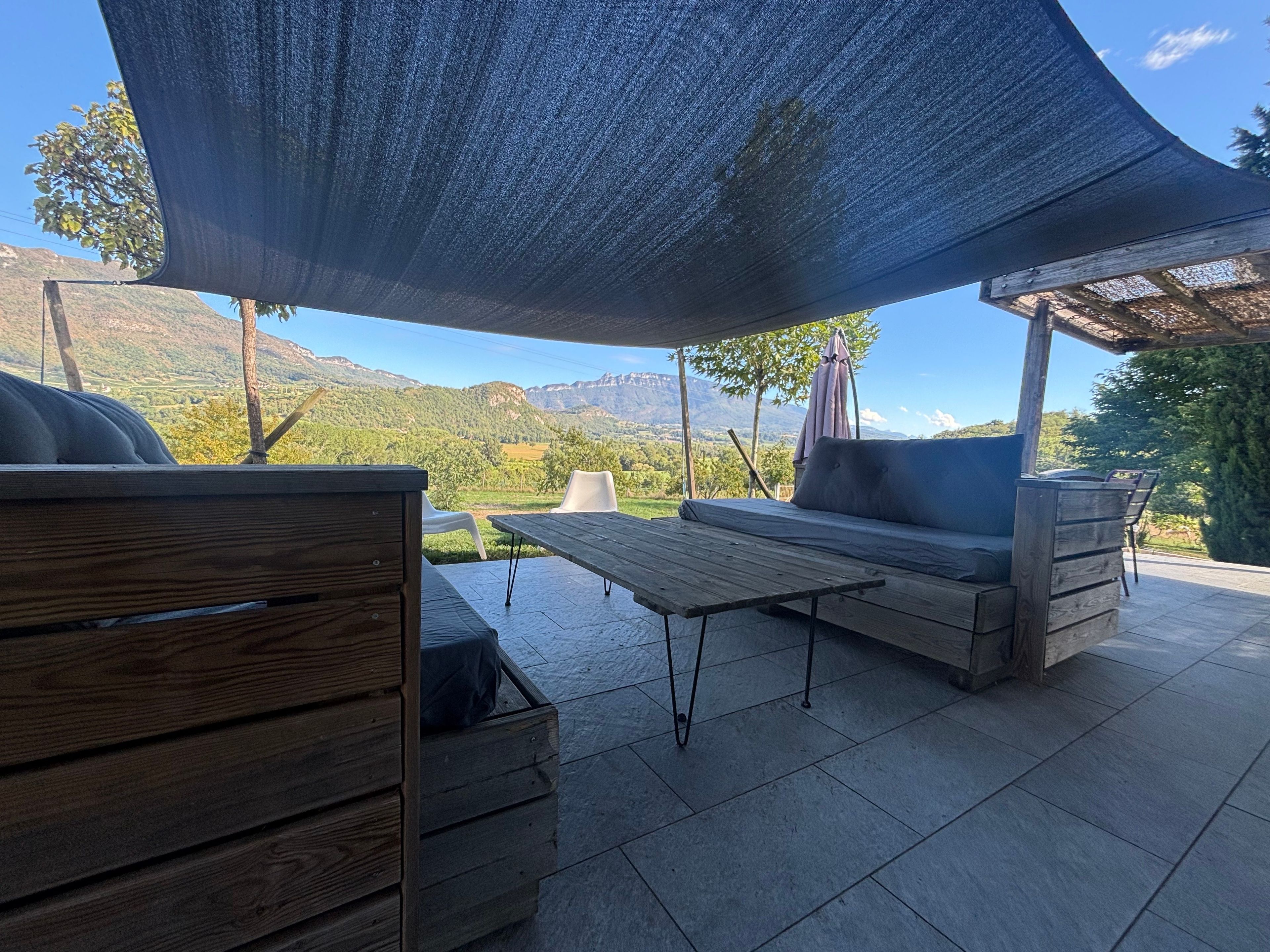 En udendørs terrasse med et træbord, to polstrede bænke og et stort skyggefuldt område.