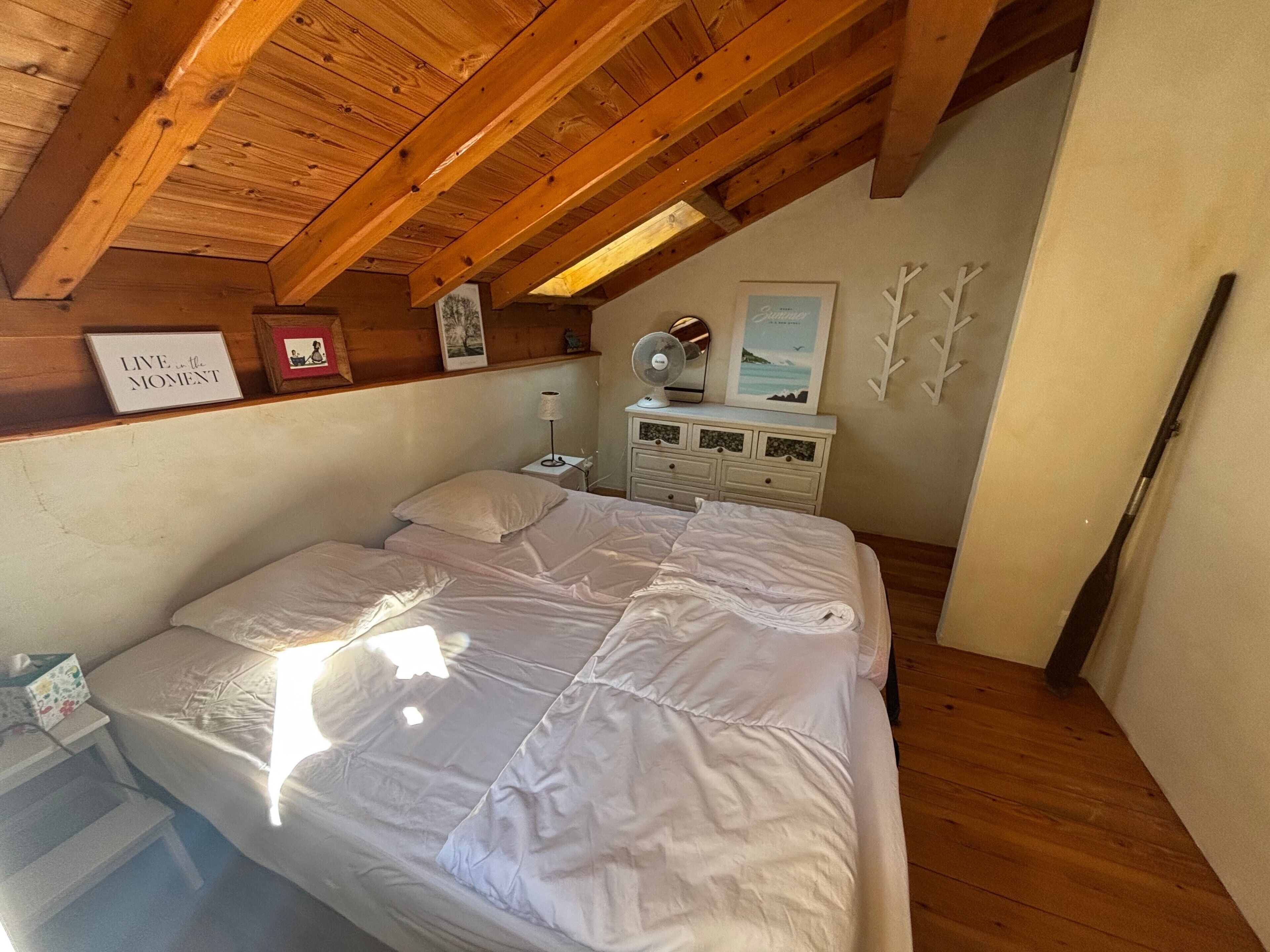 Une chambre avec un plafond en pente, un lit avec une literie blanche, une table de chevet avec une lampe et une commode avec des tiroirs.