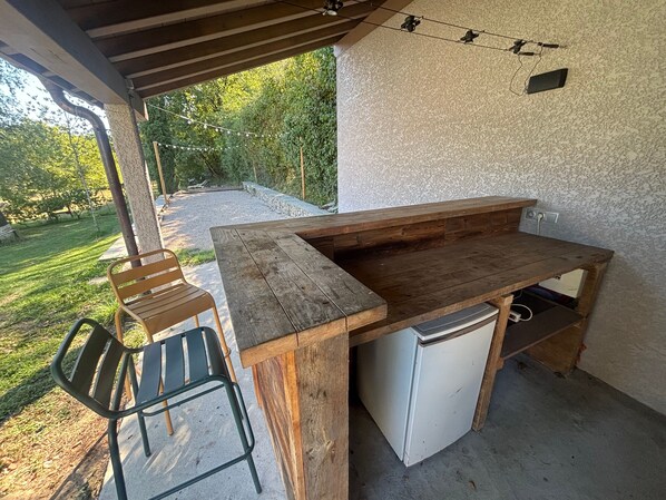 Un área de cocina al aire libre con una mesa de madera, un refrigerador empotrado y un banco.