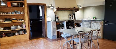 A rustic kitchen with a dining table and chairs, wooden shelves with kitchenware, and a black refrigerator.