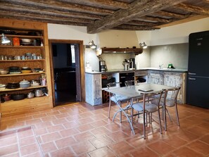 A rustic kitchen with a dining table and chairs, wooden shelves with kitchenware, and a black refrigerator. - Charming 135 m² Country House, Quiet Setting, Enclosed Garden (Chirassimont)