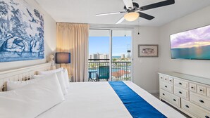 Quarto de hotel com uma cama grande, ventoinha no teto, vista para o mar e um quadro na parede.