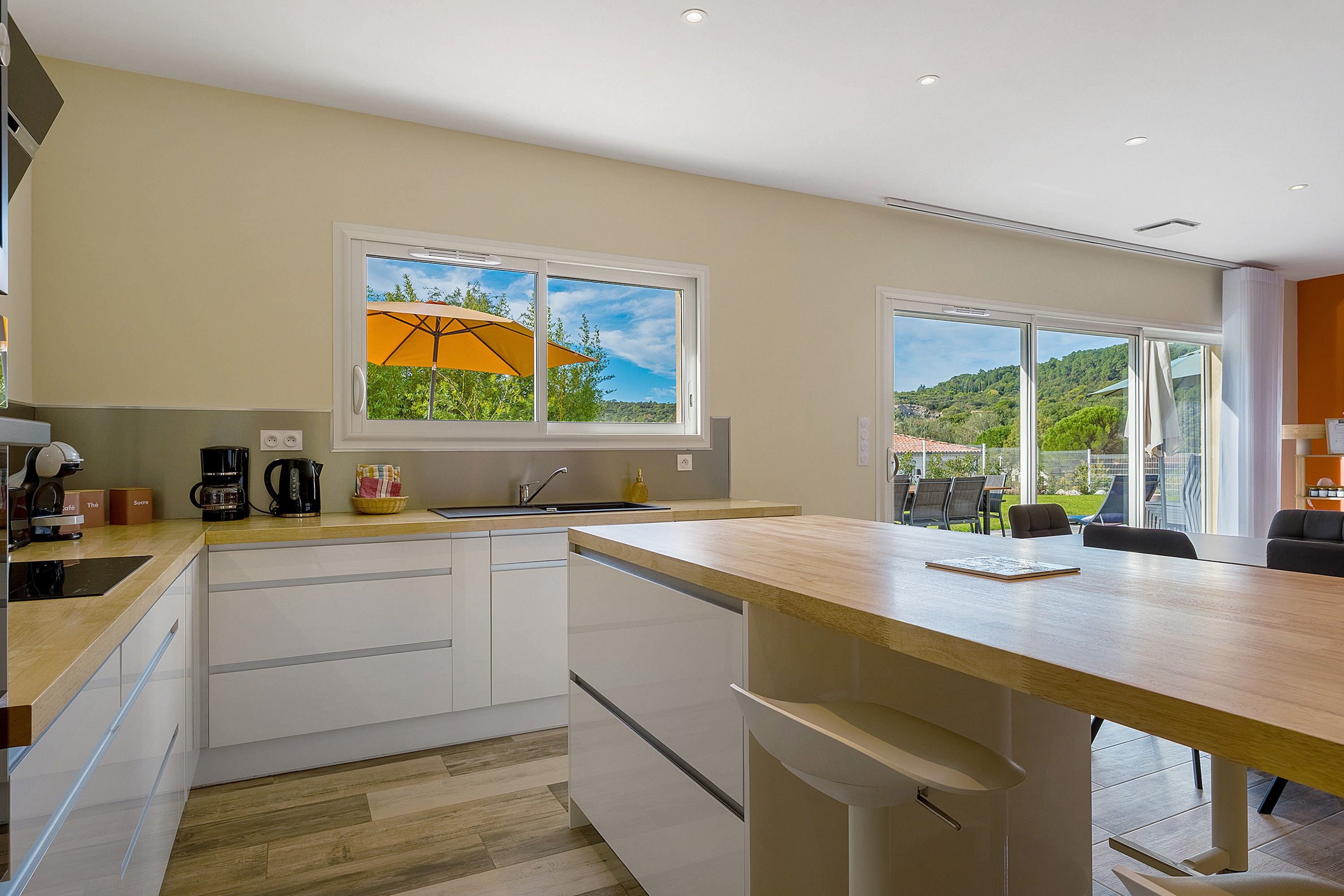 A modern kitchen with a large window, a dining area with a table and chairs, and a view of a green landscape.