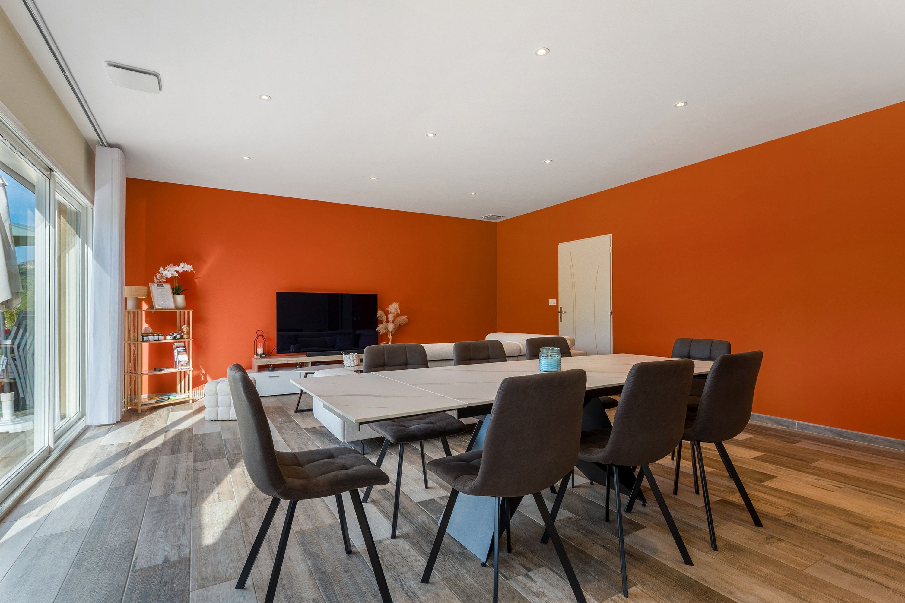 A modern dining area with a large table and chairs, a television, and a bookshelf with books and decorative items.