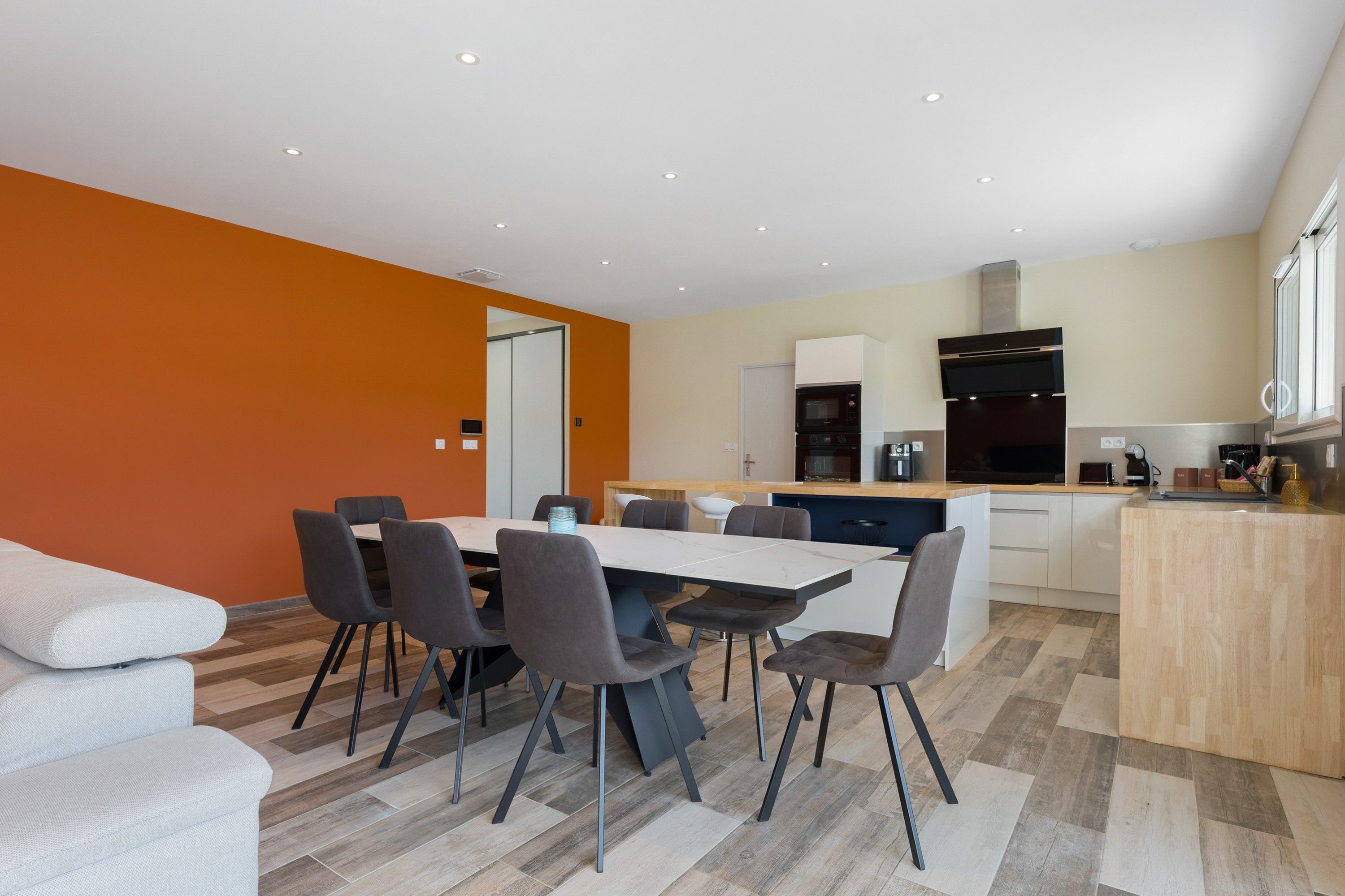 Une cuisine moderne dotée d’appareils électroménagers intégrés, un coin repas avec une grande table et des chaises, et un mur d’accent d’un orange vif.