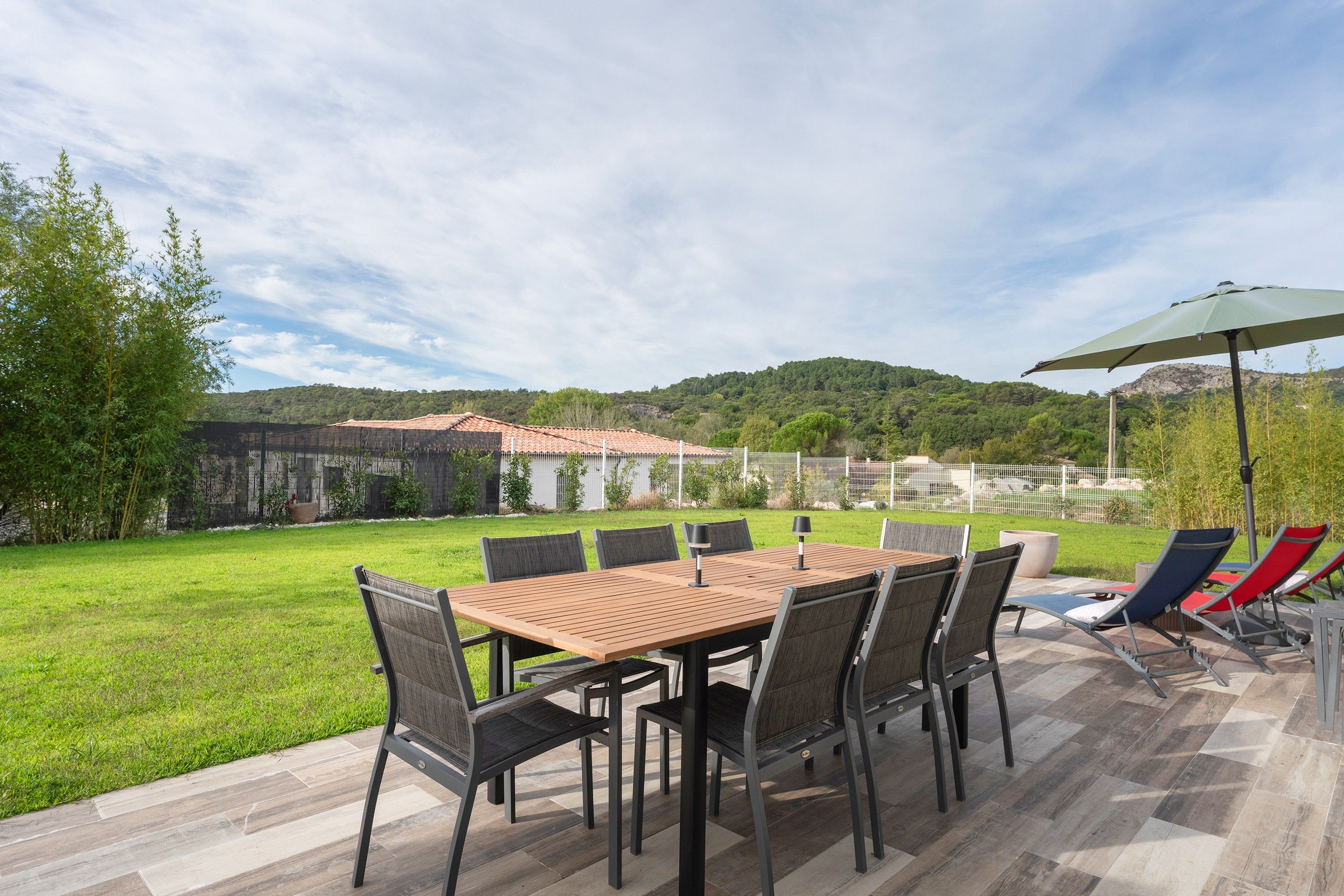 An outdoor patio with a wooden table and chairs, surrounded by a grassy area and a view of a hill.