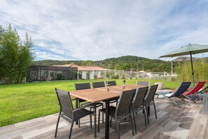 An outdoor patio with a wooden table and chairs, surrounded by a grassy area and a view of a hill.