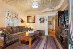 Ein Wohnzimmer mit einem gemusterten Sofa, einem Holztisch und einer dekorativen Lampe. An der Wand hängt ein gerahmtes Bild, und es gibt einen Schrank mit Glastüren.