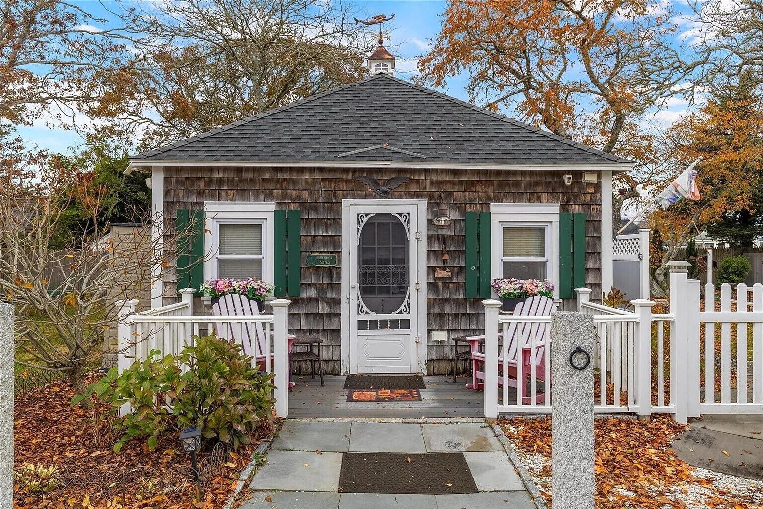 A small wooden cottage with a front porch, two white chairs, and a white picket fence.