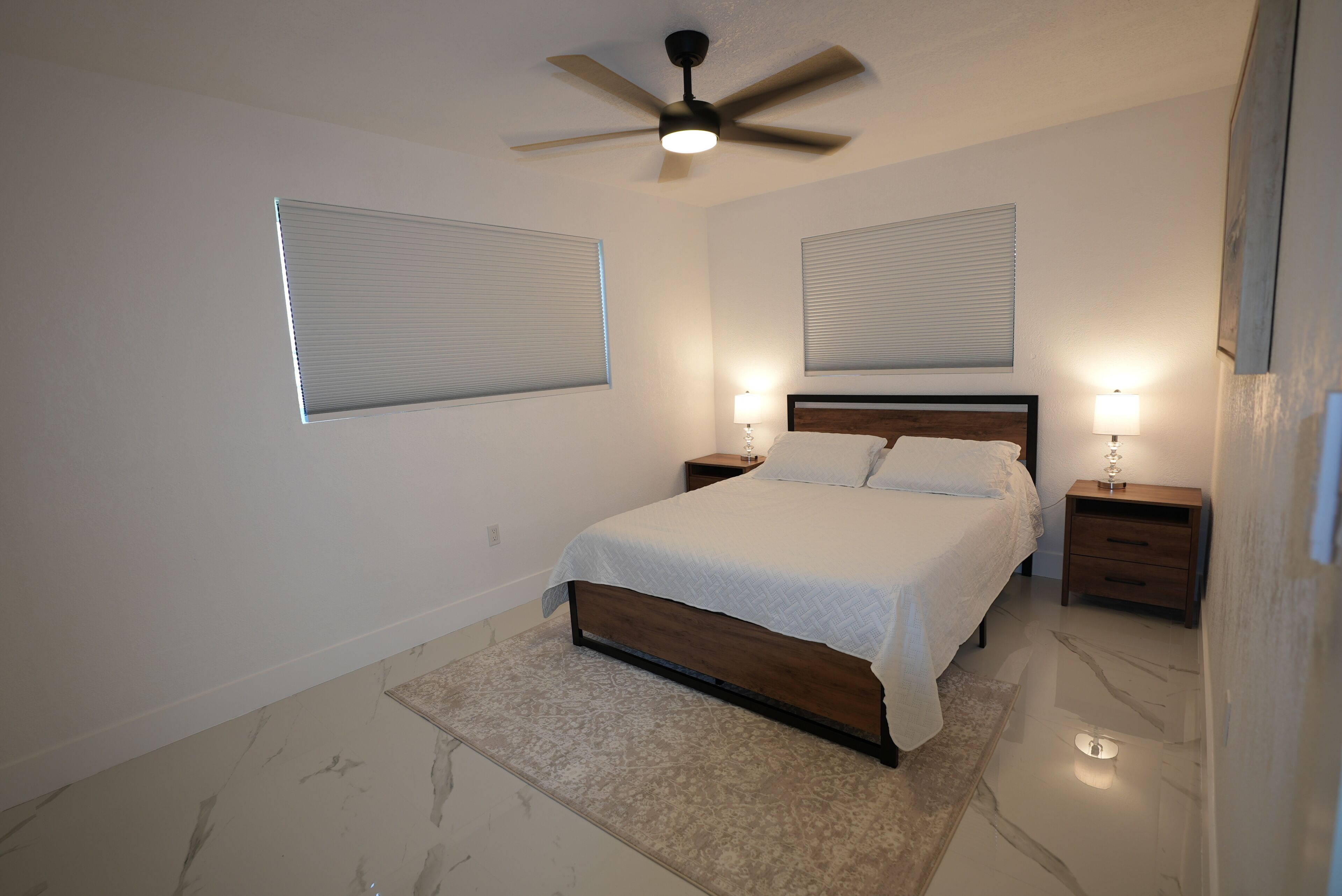 Un dormitorio con una cama, dos mesitas de noche, un ventilador de techo y dos ventanas con persianas.