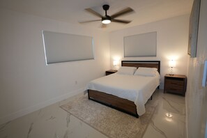 A bedroom with a bed, two bedside tables, a ceiling fan, and two windows with blinds.
