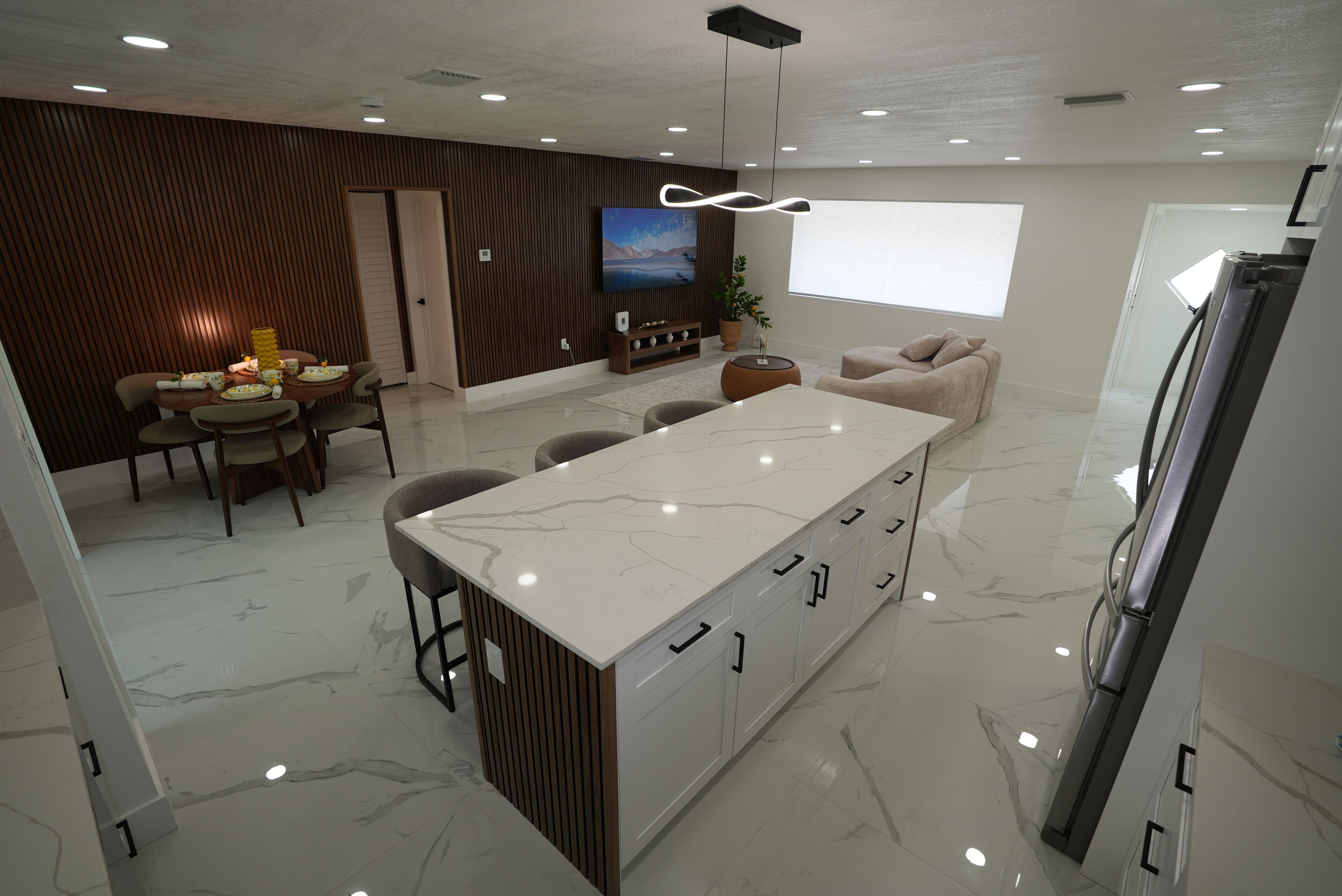 Una cocina moderna con encimera de mármol, comedor con mesa y sillas, sofá y televisor montado en la pared.
