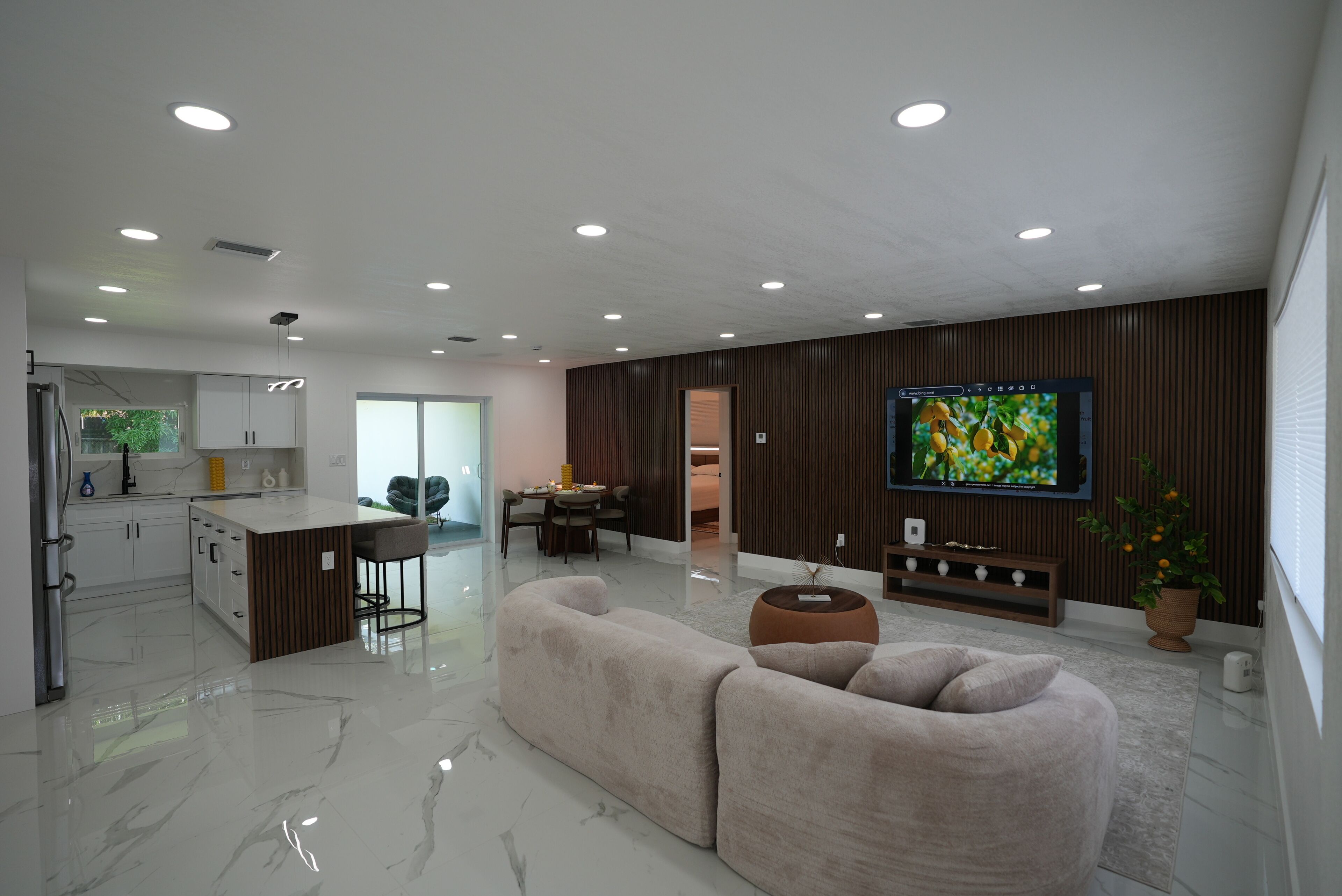 A modern living room with a sofa, a dining area, a kitchen, and a TV mounted on the wall.