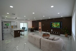 A modern living room with a sofa, a dining area, a kitchen, and a TV mounted on the wall.