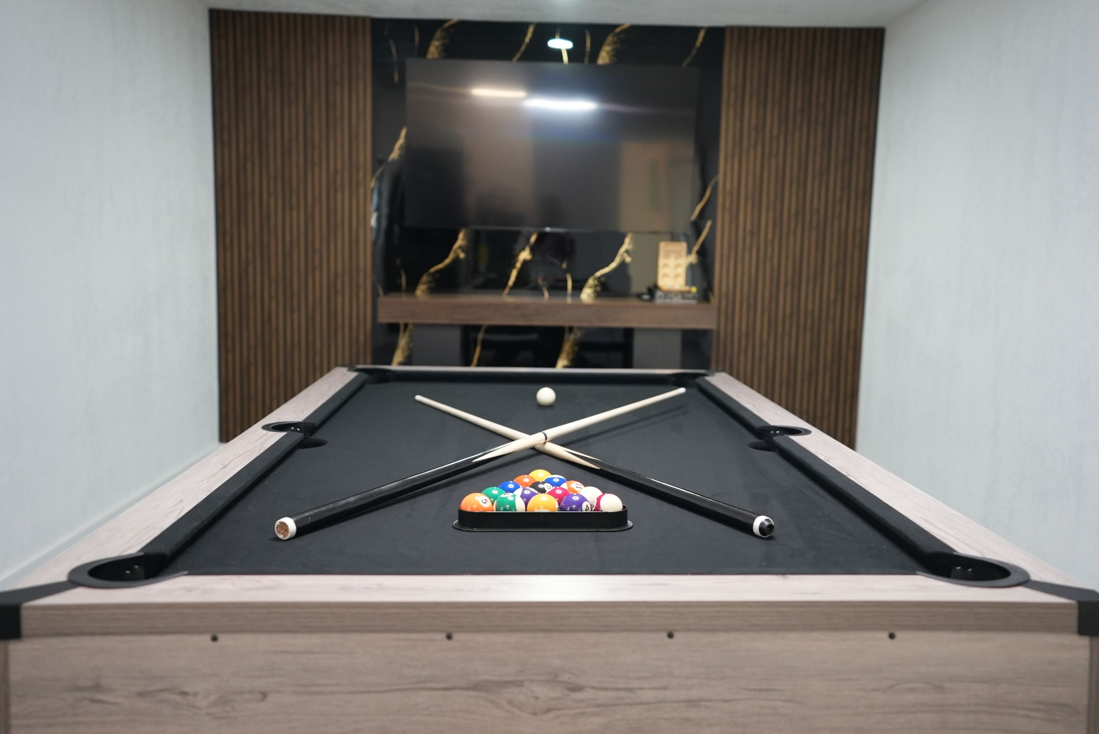 Una mesa de billar con bolas y tacos, un borde de madera y una ventana con cortinas.