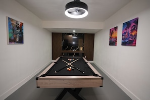 A pool table with cue sticks and balls, flanked by two framed artworks, under a ceiling light fixture.