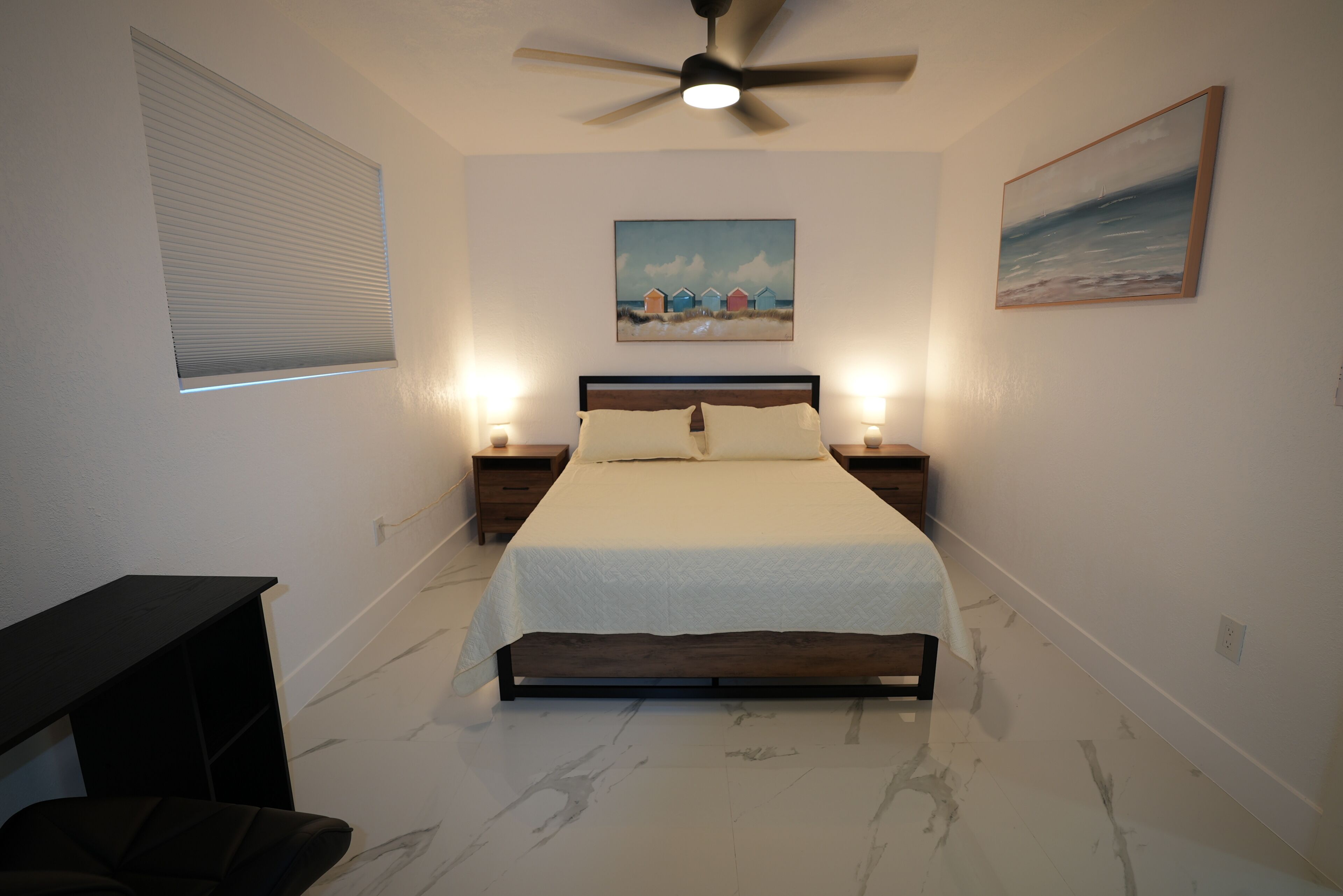 A bedroom with a bed, nightstands, a ceiling fan, a painting, and a window with blinds.