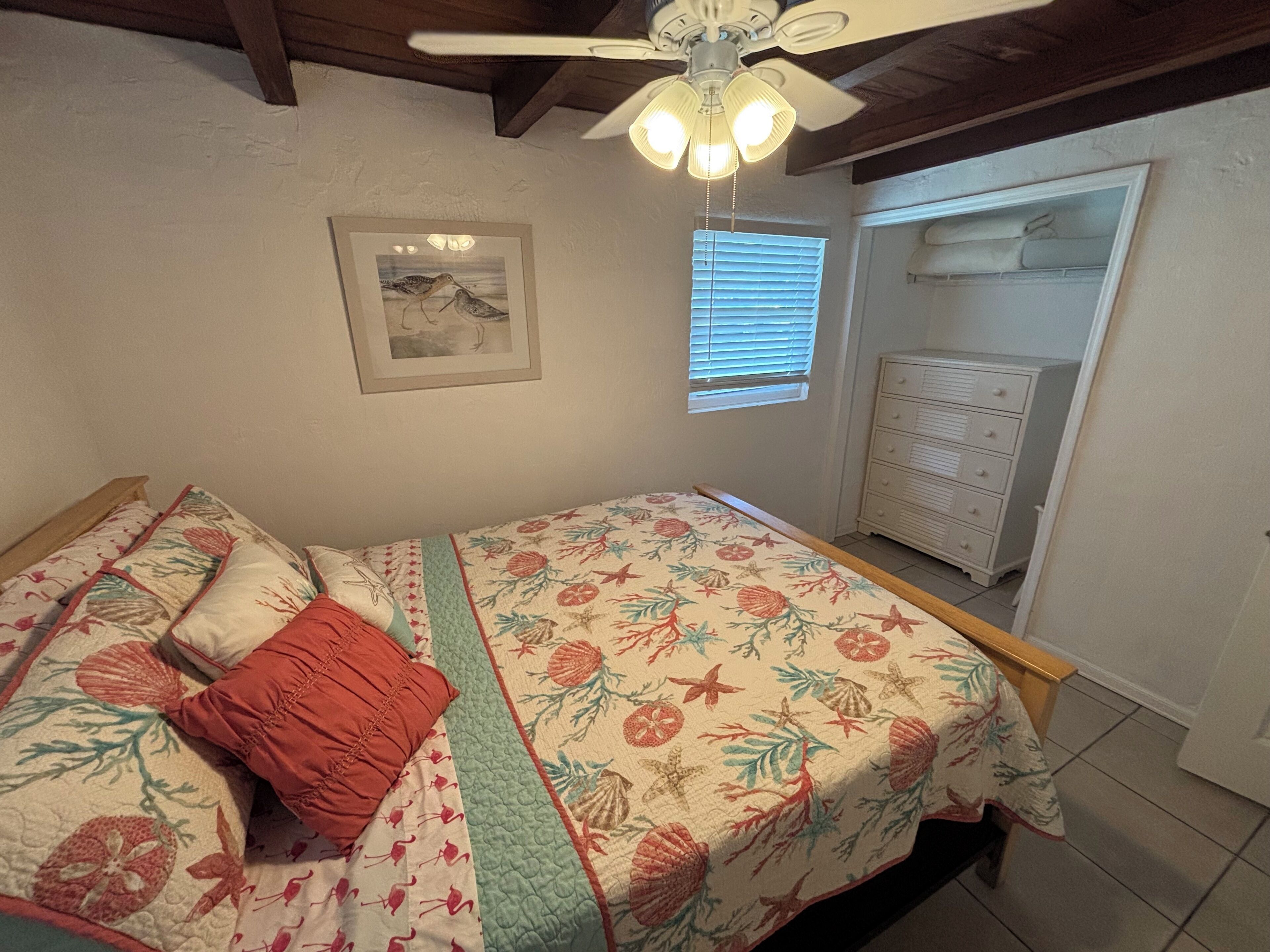 Un dormitorio con cama, ventilador de techo, ventana con persianas, cómoda y un cuadro en la pared.