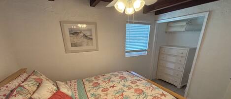 Un dormitorio con cama, ventilador de techo, ventana con persianas, cómoda y un cuadro en la pared.