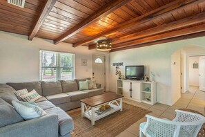 A living room with a wooden ceiling, a sofa, a coffee table, a TV, and a white cabinet. - Walk to Mashes Sands Beach, boat from your private dock, and bask in Old Florida in this one-story canal-front home (Panacea)