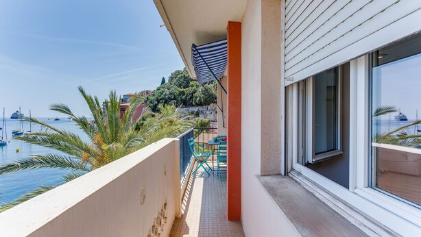 A balcony with a view of the sea and boats, a tiled floor, and a window with blinds.