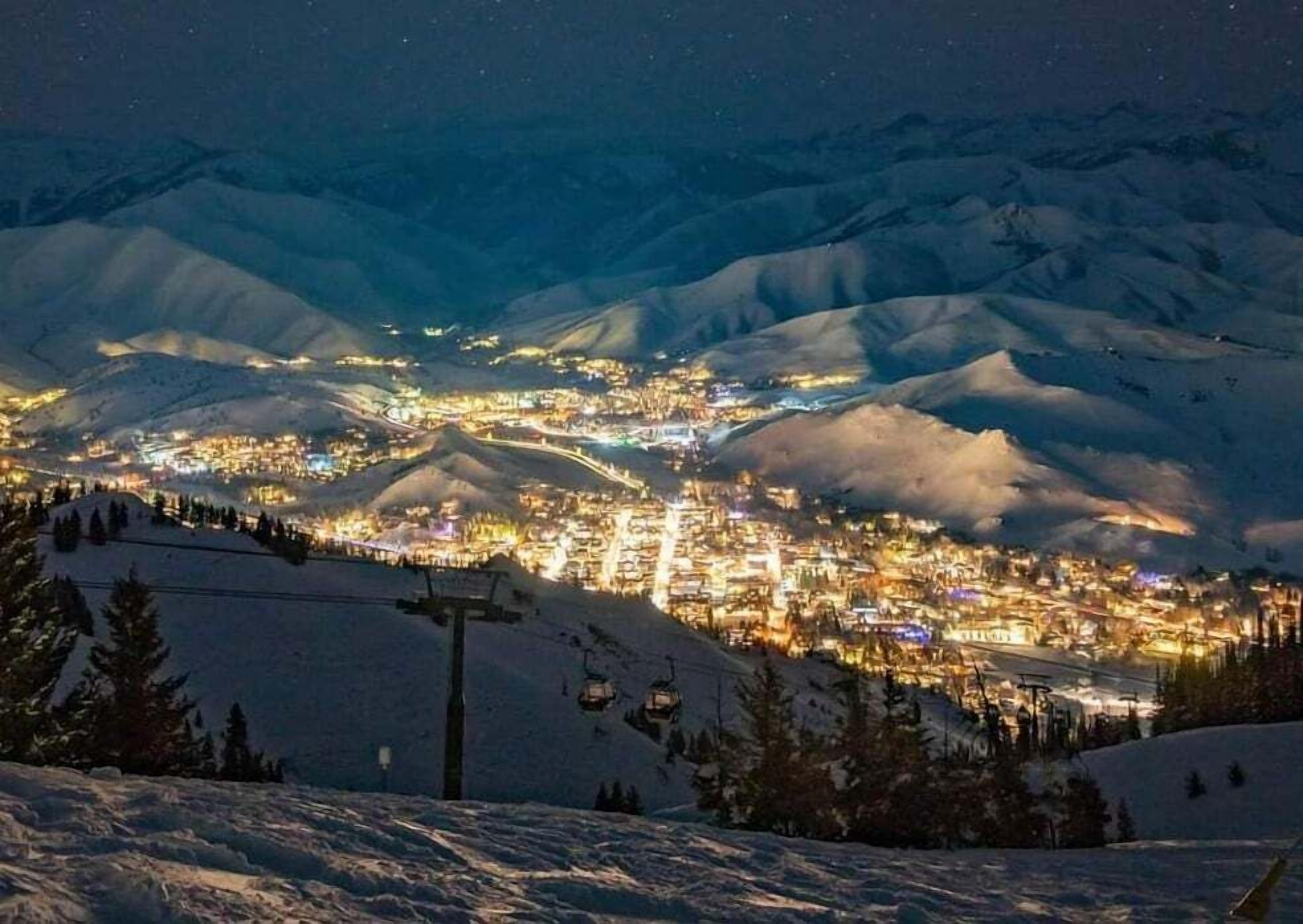 A snow-covered mountain town at night with illuminated buildings and a ski lift in the foreground.