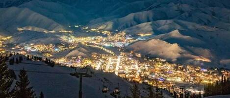 Uma cidade nas montanhas coberta de neve à noite, com edifícios iluminados e um teleférico em primeiro plano.