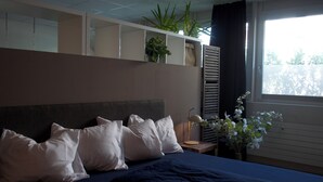 Hotellihuone, jossa on sänky, tyynyjä, yöpöytä lampun kanssa, maljakko kukkineen ja ikkuna, jossa on verhot.