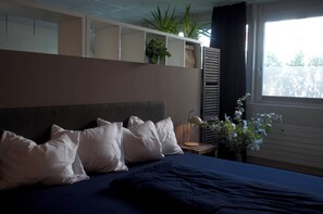 Ein Hotelzimmer mit Bett, Kissen, einem Nachttisch mit Lampe, einer Vase mit Blumen und einem Fenster mit Vorhängen.
