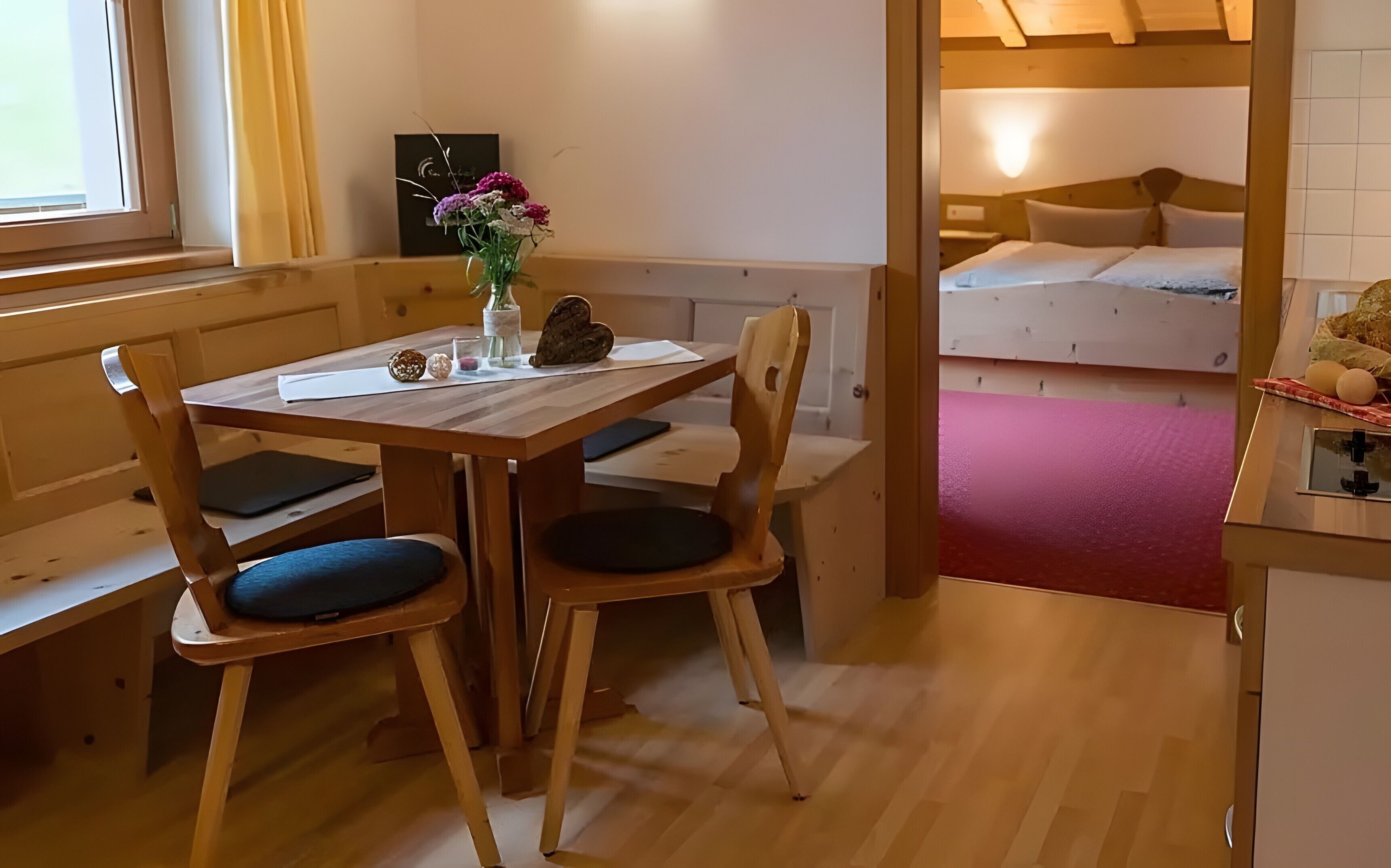 Een houten eettafel met stoelen, een vaas met bloemen en een bank in een kamer met een stapelbed op de achtergrond.